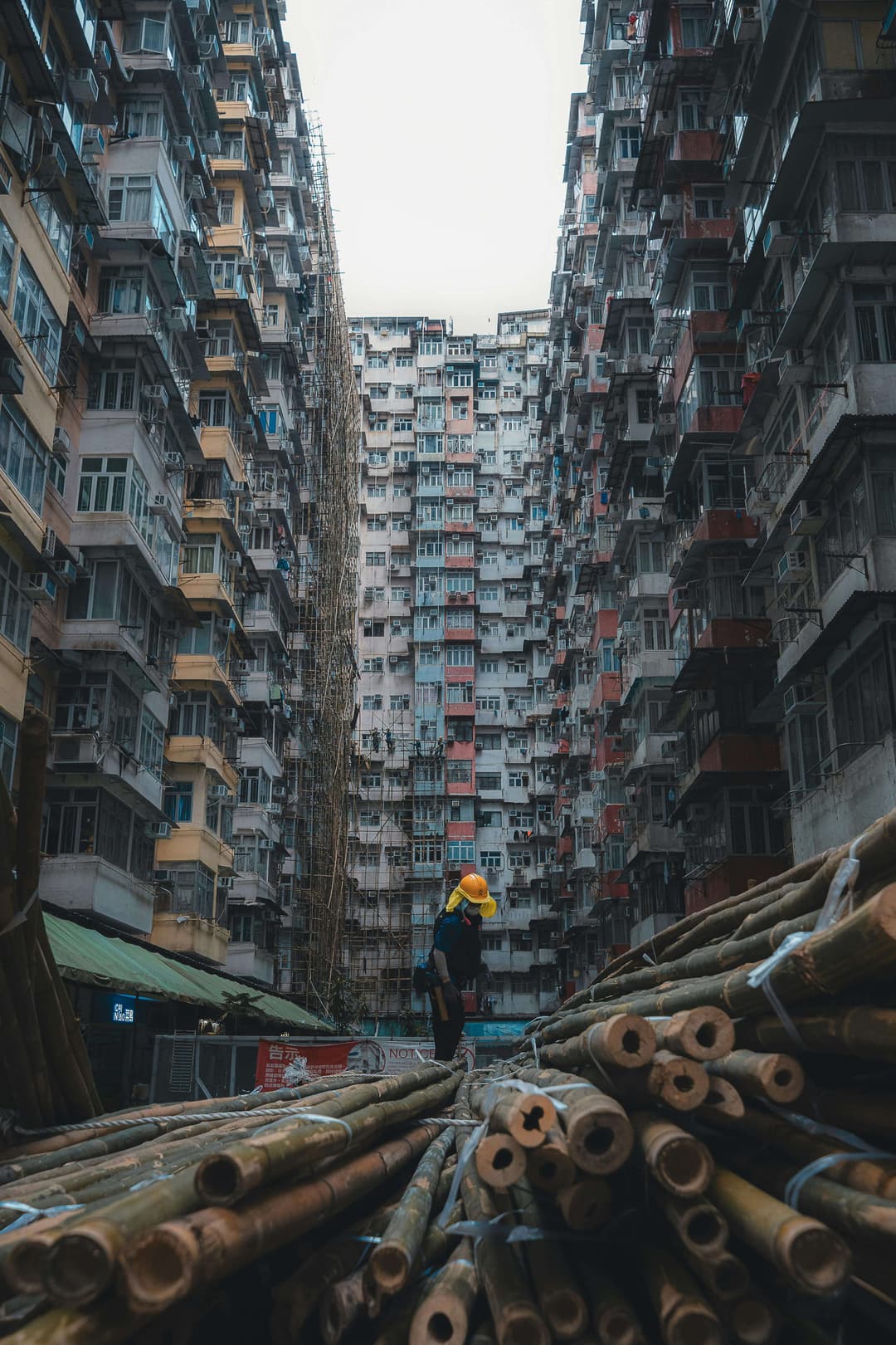 A worker stands on bamboo scaffolding in a narrow alley between tall, densely packed residential buildings.